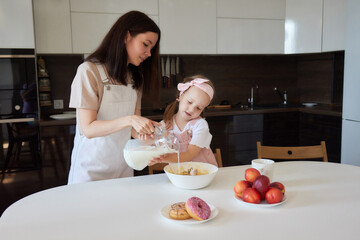 Mother and daughter preparing a sweet cake using flour, milk, sitting on chairs at a table in a...