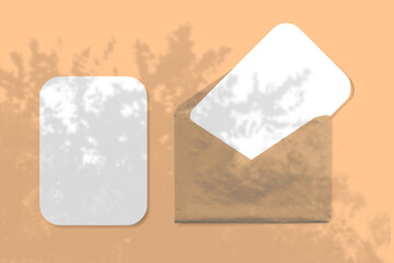 An envelope with two sheets of textured white paper on orange background. Mock up with an overlay of palm tree shadows. Natural light casts shadows from the leaves of a tree branch. Horizontal