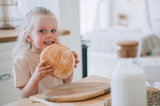 Little Swedish girl in casual holds bread smiles stands at kitchen table. Healthy food, children feeding. Childhood, healthy kids. Preschooler girl at kitchen. Food and kids. Gluten free products