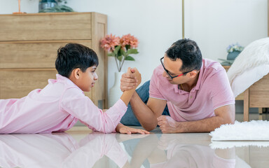 Indian happy father and son playing together, laying down on floor, doing arm wrestle with amusement and fun while staying in living room at cozy home in weekend. Education, Family, Activity Concept.