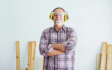 Senior Caucasian retired old happy male woodworker smiling, wearing check shirt, earmuff to protect...
