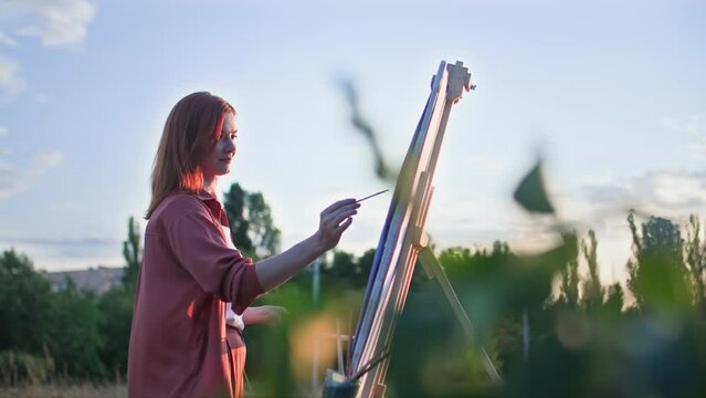 talented female artist paints a picture on canvas with paints and a brush using an easel backdrop of trees and setting sun