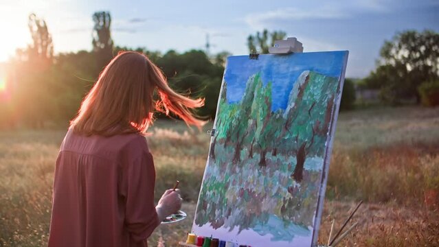 artistic hobby, a talented young woman with paints and brushes draws a creative picture using an easel in the park against the backdrop of sunlight