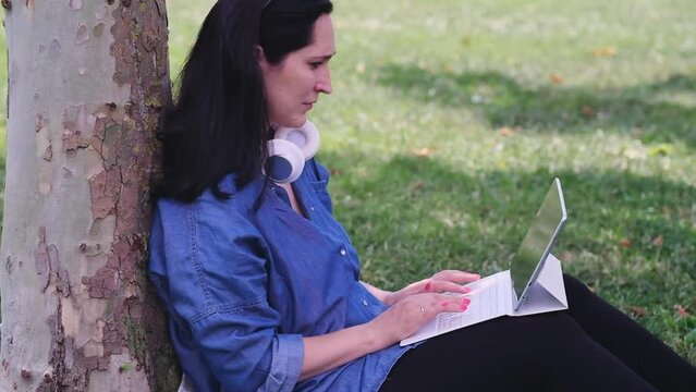 Positive woman with cordless earphones writing article on tablet computer sitting on lawn near tree in green garden freelance employee working on internet outdoors