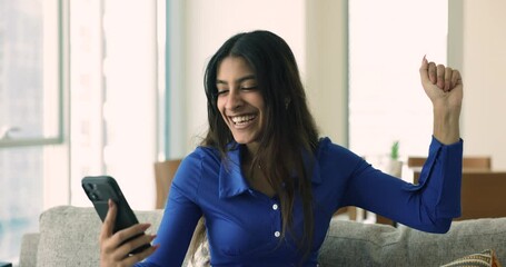 Cheerful young woman sits on sofa hold smartphone, read sms, get sell-out commercial offer, get hired, bet on-line game win. Overjoyed female celebrates lottery victory, relish success and achievement