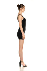 Profile Elegance: Side View Full-Length Portrait of a Stylish Young Woman in a Short Black Dress, High Heels, and Bun Hairstyle on White Background