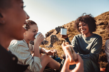 Young and diverse group of female friends taking a break from hiking and having warm drinks and snacks in the mountains