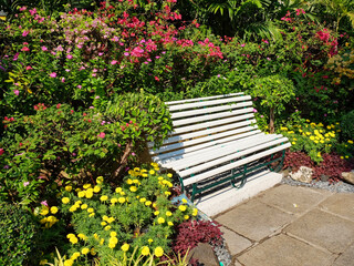 A white wooden bench sits on a stone path in a garden, surrounded by vibrant pink, red, and yellow...