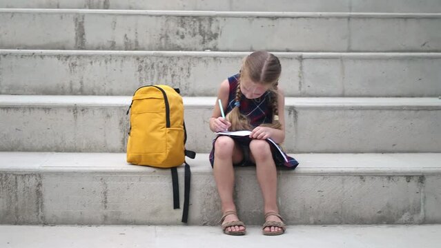 Concentrated little girl writes in exercise book sitting by schoolbag on stone steps schoolgirl with plaits does homework in school yard primary school pupil