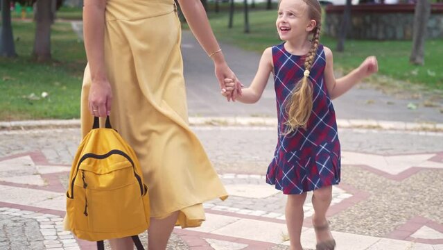 Active little girl hops walking with mother carrying backpack across green park woman in yellow dress takes playful daughter to school in garden family relations