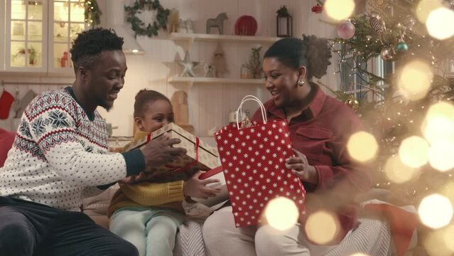 Medium shot of joyful African American family of three sitting on couch by Christmas tree exchanging presents with each other