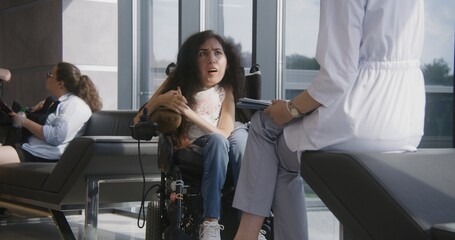 Woman with SMA in electric wheelchair talks to doctor in hospital or clinic lobby. Female physician consults patient with physical disability. People sit on sofa in modern medical center waiting area.