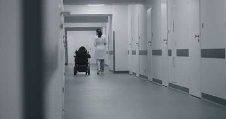 Female doctor walks down the clinic hallway, consults woman with physical disability. Physician talks about medical procedures with patient in electric wheelchair. Corridor of modern medical facility.