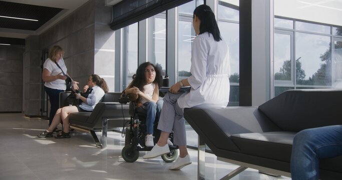 Woman with disability in motorized wheelchair talks to doctor in modern hospital or clinic lobby. Professional physician sits on sofa, consults female patient with spinal muscular atrophy. Dolly shot.