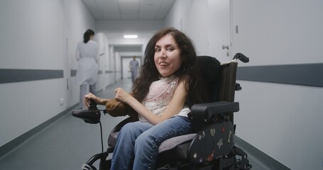 Woman with spinal muscular atrophy in motorized wheelchair in the middle of clinic corridor smiles...