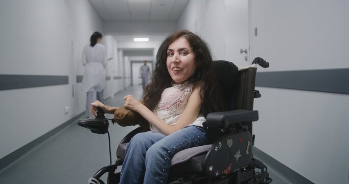 Woman with spinal muscular atrophy in motorized wheelchair in the middle of clinic corridor smiles and looks at camera. Medical staff walk in hallway of hospital or medical facility in the background.