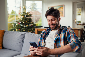 Happy young man sitting on the sofa using mobile phone during Christmas time at home