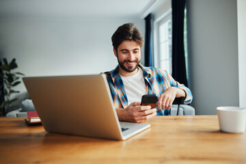 Happy young man at home using smartphone while sitting at the table working remotely