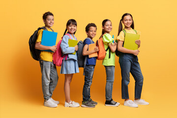 Multiracial kids posing with backpacks and books isolated on yellow