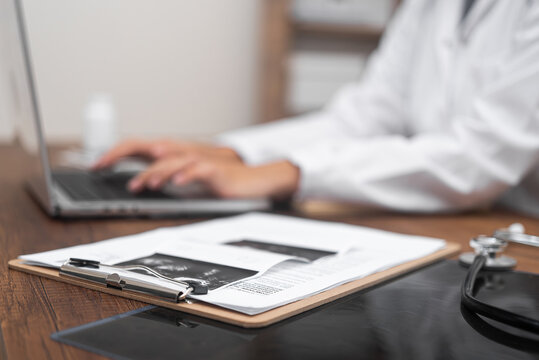 X-ray and ultrasound examination pictures lie on table against working doctor woman in white lab coat works at laptop processing results of patient tests on blurred background