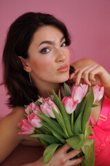 Beautiful brunette woman sitting on pink velvet sofa with a bucket of flowers tulips. Girl in pink bodysuit near window. Portrait of young woman smile. Living room.