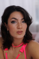 Beautiful brunette woman sitting on pink velvet sofa. Girl in pink bodysuit near window, thoughtful. Portrait of young woman smile. Living room.