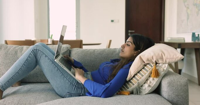 Side view young 20s Indian woman lying on couch with wireless computer, spend free time on internet, prepare task for college exam, make assignment, websurfing information, working or studying on-line