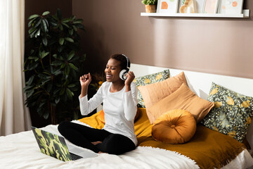 Photo of delighted young girl afro american lady sitting on bed dancing relaxing listening to music smiling sitting in fronf of computer
