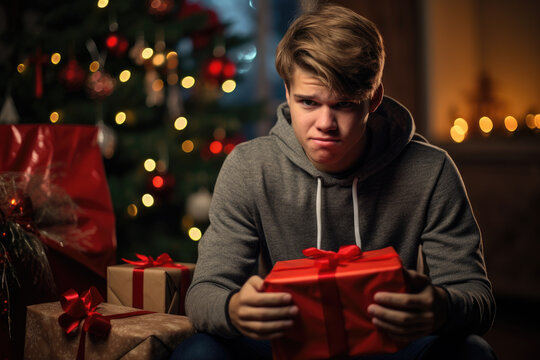 Displeased Unhappy Teenager Unsatisfied With Christmas Present. Boy Grimacing Skeptical Holding Gift Box Sitting Near Decorated Christmas Tree At Home