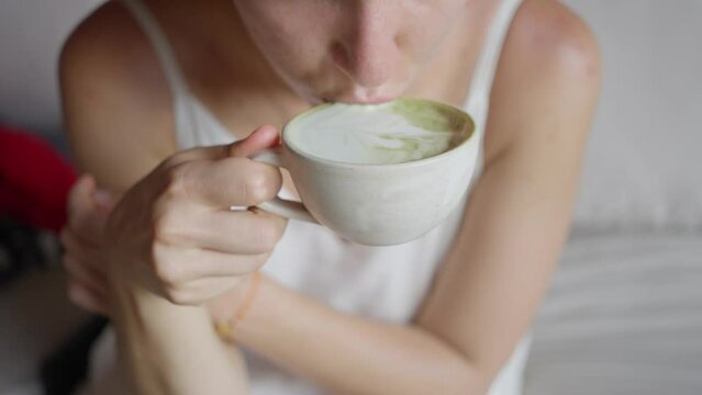 The Girl in a white dress, with a white cup, drinks a green matcha latte, slow motion. Close-up. Hot green tea drink