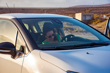 White woman in sunglasses getting into the white car preparing her seat to start driving in a dry brown mountains landscape at sunset.