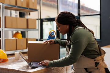 Stockroom employee checking customers orders on laptop, preparing packages using cardboard boxes in storage room. African american worker wearing industrial overall during storehouse inventory