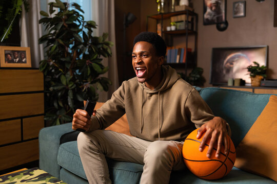 An African American basketball fan enjoys the game from the comfort of his living room, holding a ball and cheering on his team in front of the TV