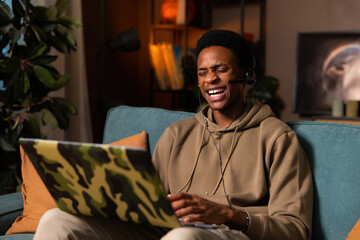 Happy dark skinned black man sitting in the living room wearing wireless headset headphones watching funny comedy film movie laughing spending time with fun at weekends relaxing after hard day - obrazy, fototapety, plakaty