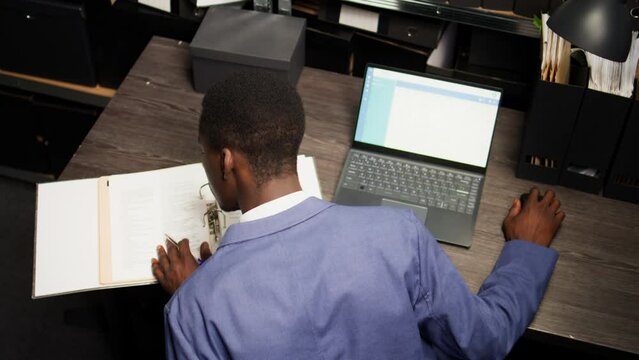 Male inspector observed diligently analyzing files and evidence acquired during research in archive room. Law officer uses laptop to cross-reference records and do a background check. Aerial shot.