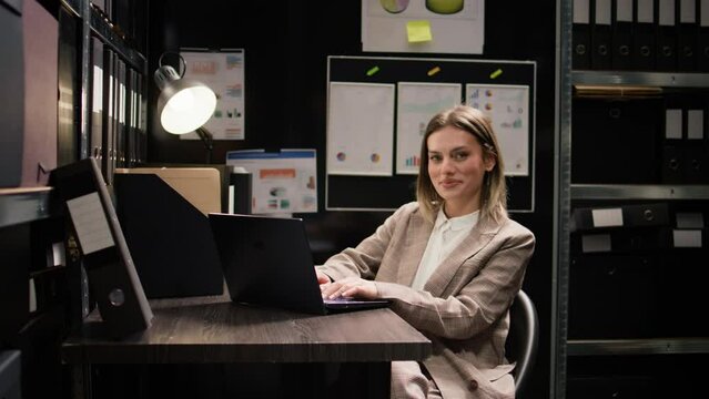 Private investigator examines evidence, looks through documents, and uses laptop in office. Portrait view of female police officer searching criminal records and files on minicomputer. Zoom-out shot.