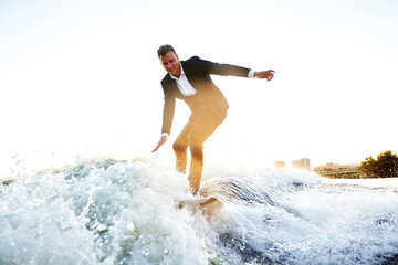 Young man in classic suit rides a wakeboard on the river or lake near city. Clerk escaped from a stuffy office to take up his favorite active sport. Best summer leisure after routine work. Sun flare.