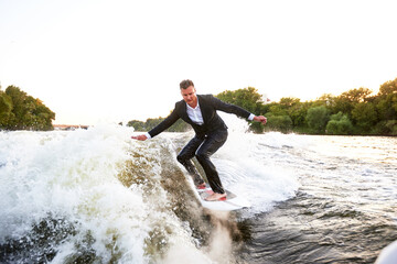 Young man in classic suit rides a wakeboard on the river or lake near city. A careless clerk escaped from a stuffy office to take up his favorite active sport. Best summer leisure after routine work.