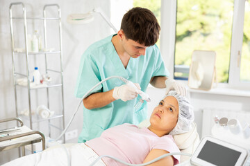 Young male cosmetologist performs hardware facial moisturizing procedure to young female patient