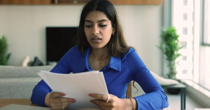 Serious Indian woman sit at desk hold documents read bad news, eviction notification, feels desperate about financial problems. Young female student worried college expulsion, failed exam test results