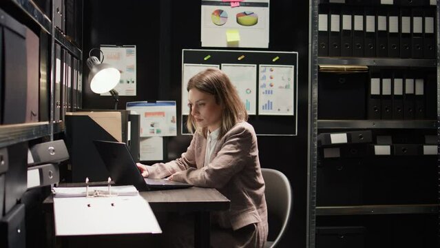 In criminal investigation office, dedicated caucasian policewoman examines evidence with personal computer. Laptop, files, and meticulous investigation all help in the hunt for clues and statements.