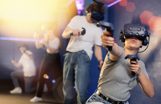 Cheerful emotional young woman in VR headset fully immersed in game, actively engaged in virtual reality world, manipulating objects or shooting targets with joysticks in hands..