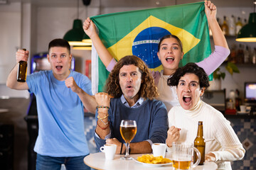 Group of friends, sports supporters cheering for favorite team with flag of Brazil while watching...