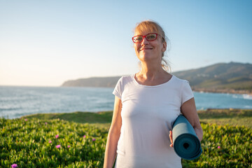 Senior woman ready for yoga exercises on seashore. Pensioner yogini holding yogamat. Mature woman going to practise yoga at sunrise at beach. Concept of healthy lifestyle on retirement