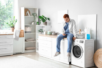 Young man with mobile phone sitting near washing machine at home