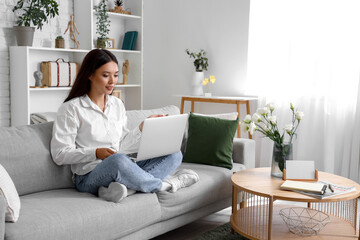 Beautiful young Asian woman with laptop sitting on sofa in living room