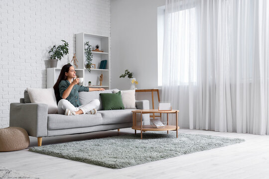 Beautiful Young Woman With Cup Of Coffee Sitting On Sofa In Living Room