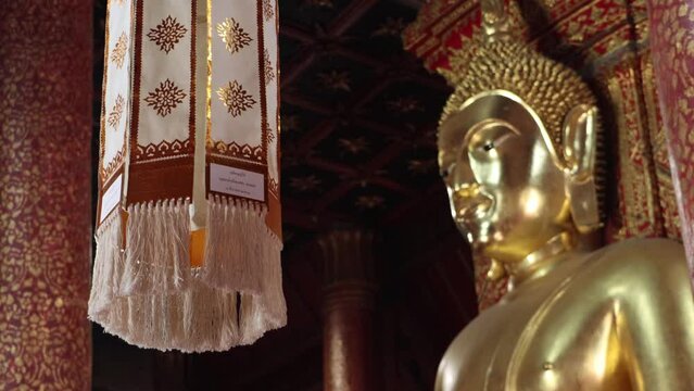 Decorative fabrics with religious text hang from the ceiling of a Buddhist temple with a gilded Buddha statue, Thailand
