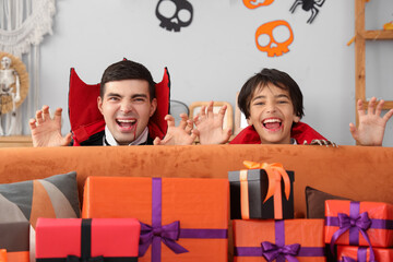 Scary little boy and his father dressed for Halloween as vampires behind sofa with gifts at home