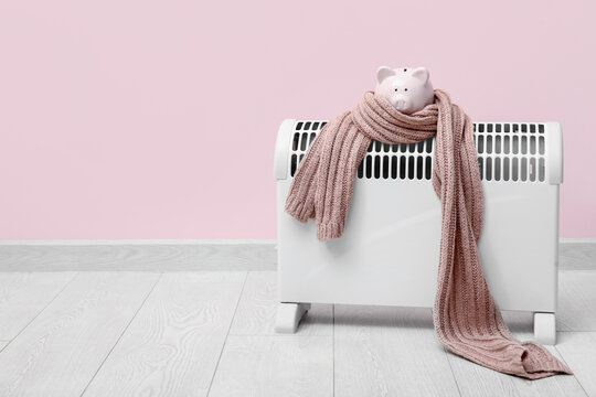 Electric convector heater, piggy bank and scarf near pink wall. Heating season